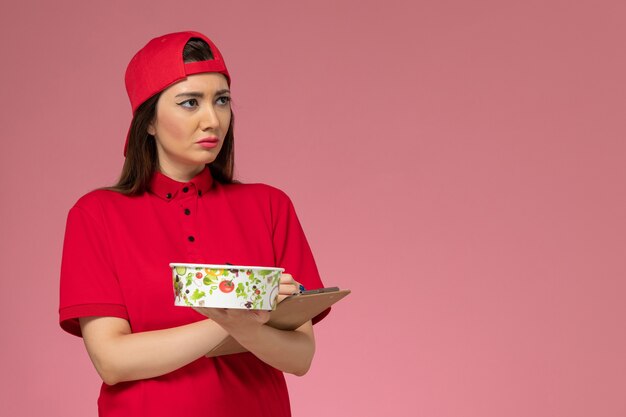 Vue de face femme courrier en cape uniforme rouge avec bloc-notes de bol de livraison ronde sur ses mains sur mur rose clair, employé de livraison d'emploi uniforme de travail
