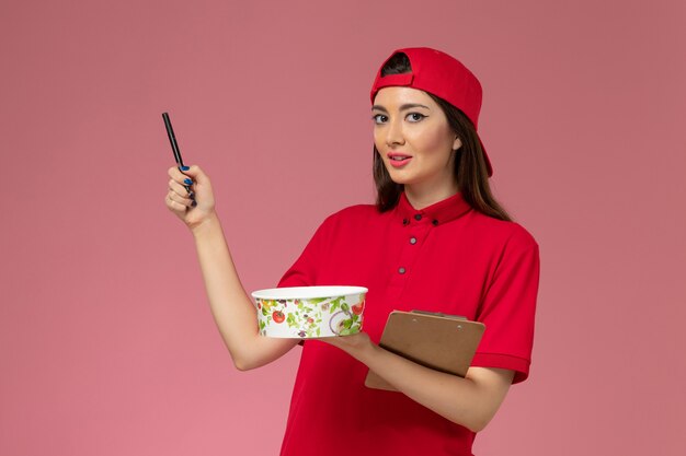 Vue de face femme courrier en cape uniforme rouge avec bloc-notes de bol de livraison ronde et stylo sur ses mains sur mur rose clair, employé de livraison uniforme