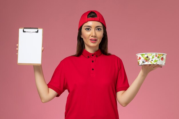 Vue de face femme courrier en cape uniforme rouge avec bol de livraison rond et bloc-notes sur ses mains sur le mur rose clair, employé de livraison uniforme
