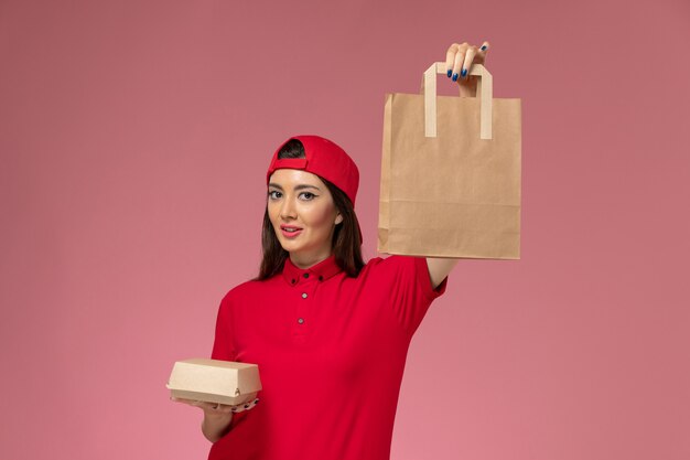 Vue de face femme courrier en cape uniforme rouge avec différents colis de livraison sur ses mains sur le mur rose, travail d'employé de livraison uniforme