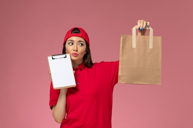 Vue de face femme courrier en cape uniforme rouge avec paquet de livraison de papier et bloc-notes pensant sur ses mains sur l'employé de livraison uniforme de bureau rose