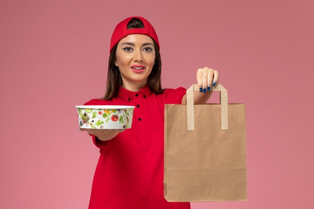 Vue de face femme courrier en cape uniforme rouge avec paquet de livraison de papier et bol sur ses mains sur le mur rose, employé de travail de livraison uniforme