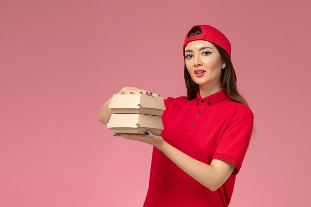 Vue de face femme courrier en cape uniforme rouge avec peu de colis alimentaires de livraison sur ses mains sur mur rose clair, employé de la prestation de services