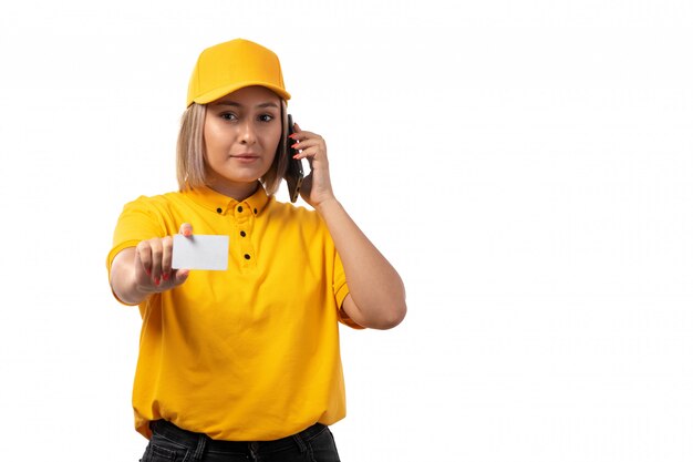 Une vue de face femme courrier en chemise jaune casquette jaune et jean noir tenant une carte blanche parler au téléphone sur blanc