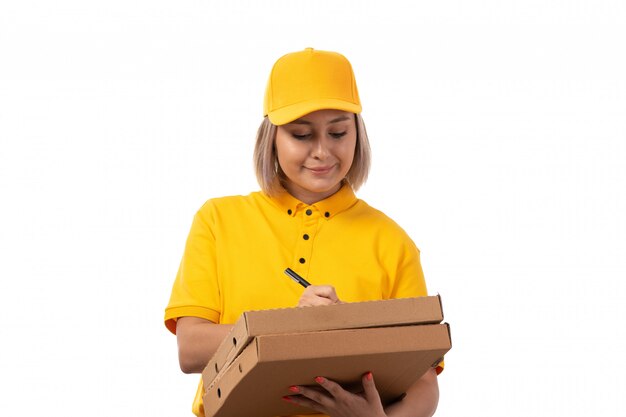 Une vue de face femme courrier en chemise jaune casquette jaune jeans noirs tenant des boîtes à pizza écrit souriant sur blanc