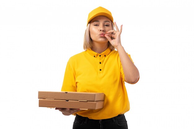 Une vue de face femme courrier en chemise jaune casquette jaune et jeans noirs tenant des boîtes à pizza montrant un délicieux geste sur blanc
