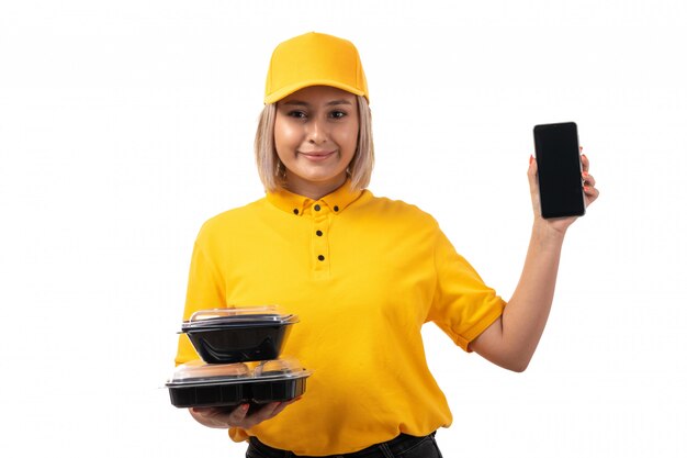 Une vue de face femme courrier en chemise jaune casquette jaune et jeans noirs tenant des bols avec de la nourriture et smartphone souriant sur blanc