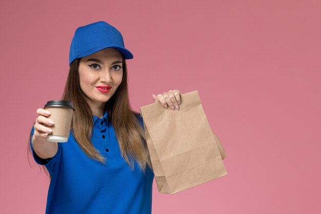 Vue de face femme courrier en uniforme bleu et cape tenant la livraison tasse de café paquet alimentaire sur le mur rose