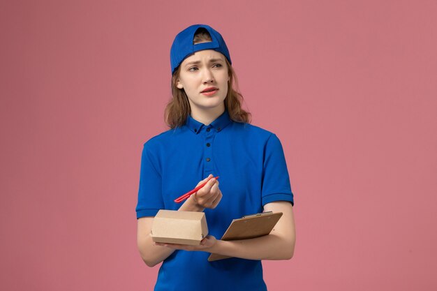 Vue de face femme courrier en uniforme bleu et cape tenant peu de bloc-notes de colis de nourriture de livraison et d'écriture sur le mur rose, le travail des employés du service de livraison