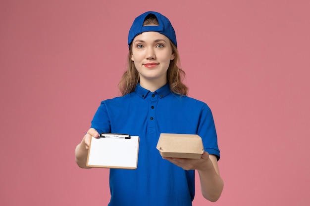 Vue de face femme courrier en uniforme bleu et cape tenant peu de colis de nourriture de livraison avec bloc-notes sur mur rose, employé du service de livraison d'emploi