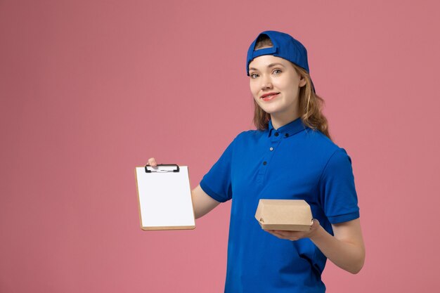 Vue de face femme courrier en uniforme bleu et cape tenant peu de colis de nourriture de livraison avec bloc-notes sur mur rose, employé du service de livraison d'emploi