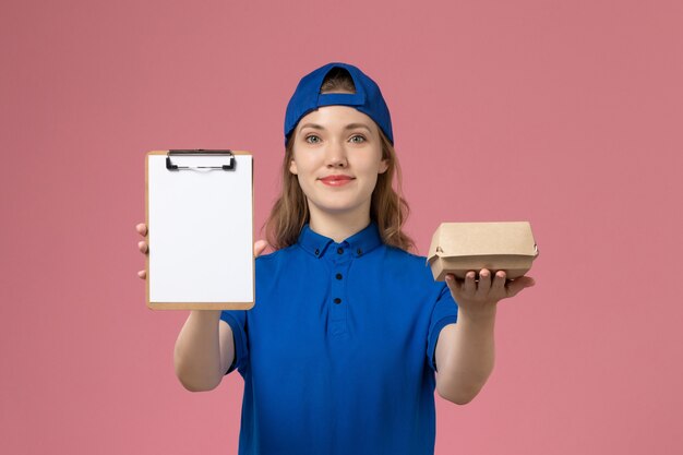 Vue de face femme courrier en uniforme bleu et cape tenant peu de colis de nourriture de livraison et bloc-notes sur le mur rose, employé du service de livraison