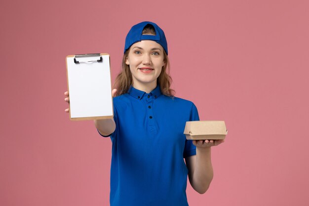 Vue de face femme courrier en uniforme bleu et cape tenant peu de colis de nourriture de livraison et bloc-notes sur mur rose, employé du service de livraison