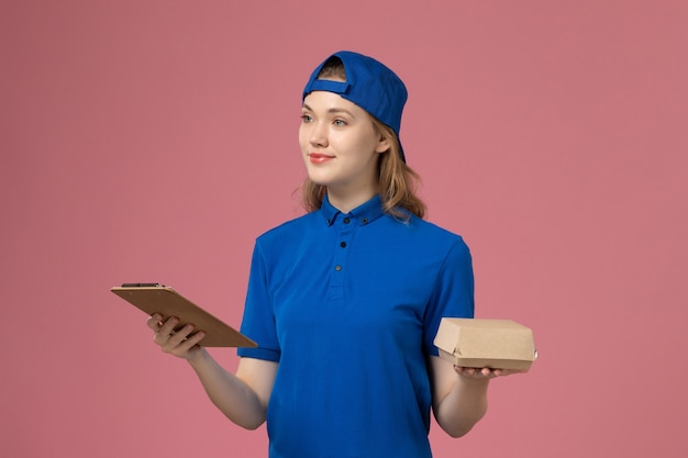 Vue de face femme courrier en uniforme bleu et cape tenant peu de colis de nourriture de livraison et bloc-notes sur mur rose, employé du service de livraison