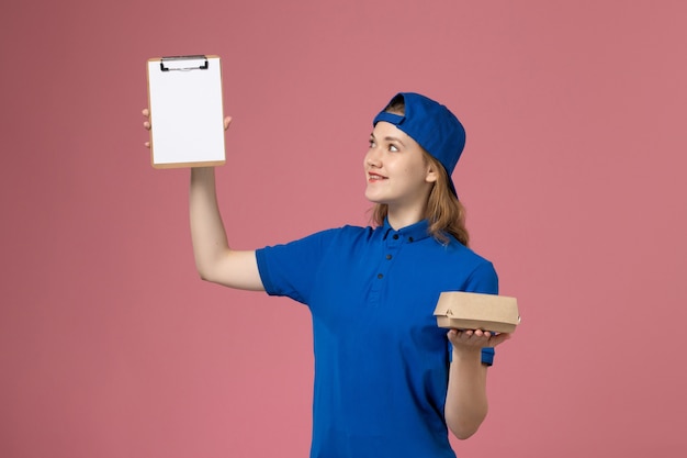 Vue de face femme courrier en uniforme bleu et cape tenant peu de colis de nourriture de livraison et bloc-notes sur mur rose, travail des employés du service de livraison