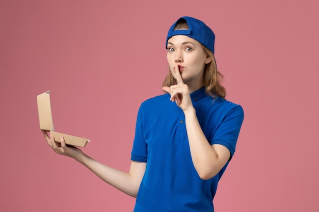 Vue de face femme courrier en uniforme bleu et cape tenant peu de colis de nourriture de livraison sur un mur rose clair, fille d'un employé de service uniforme de livraison