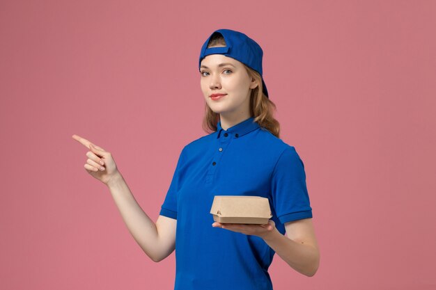 Vue de face femme courrier en uniforme bleu et cape tenant peu de colis de nourriture de livraison sur le mur rose, entreprise de service uniforme travail travailleur fille