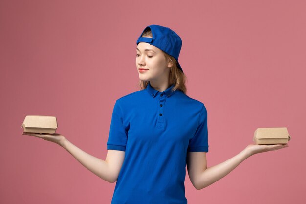 Vue de face femme courrier en uniforme bleu et cape tenant peu de colis de nourriture de livraison sur le mur rose, entreprise de service uniforme de travail