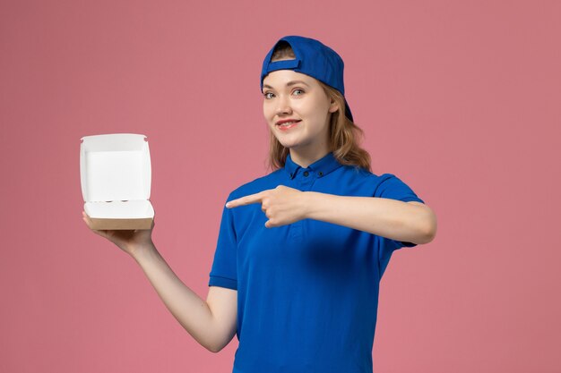 Vue de face femme courrier en uniforme bleu et cape tenant peu de colis de nourriture de livraison sur le mur rose, travail d'un employé de service uniforme de livraison