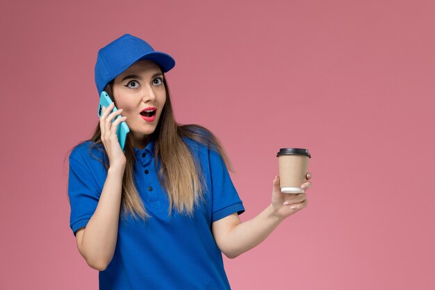 Vue de face femme courrier en uniforme bleu et cape tenant la tasse de café de livraison à l'aide d'un téléphone sur mur rose