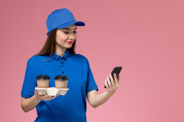 Vue de face femme courrier en uniforme bleu et cape tenant des tasses de café de livraison à l'aide de son téléphone sur mur rose