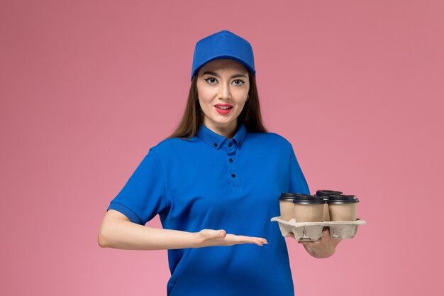 Vue de face femme courrier en uniforme bleu et cape tenant des tasses de café de livraison sur un bureau rose clair