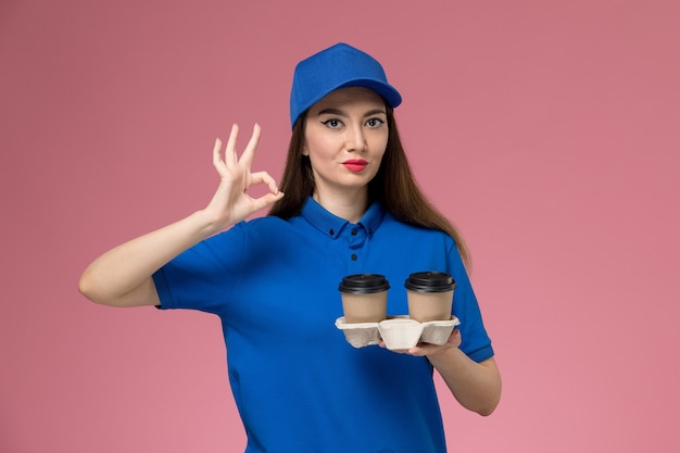 Vue de face femme courrier en uniforme bleu et cape tenant des tasses de café de livraison sur le bureau rose