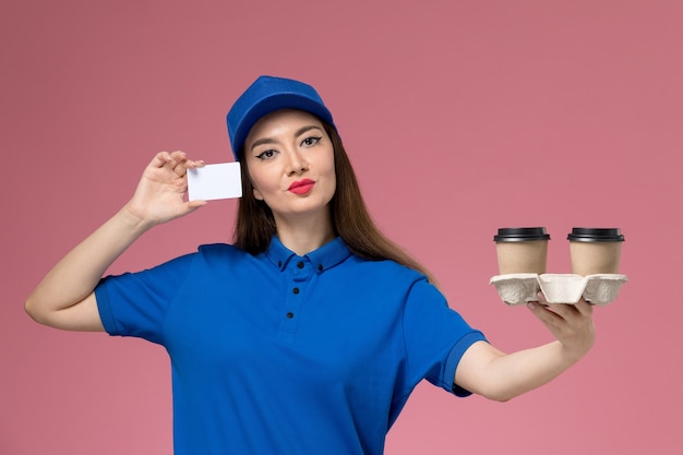 Vue de face femme courrier en uniforme bleu et cape tenant des tasses de café de livraison marron et carte posant sur mur rose