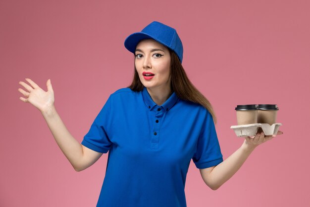 Vue de face femme courrier en uniforme bleu et cape tenant des tasses de café de livraison sur le mur rose travailleur de l'emploi