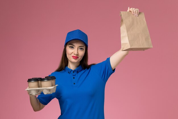 Vue de face femme courrier en uniforme bleu et cape tenant des tasses à café paquet alimentaire sur mur rose