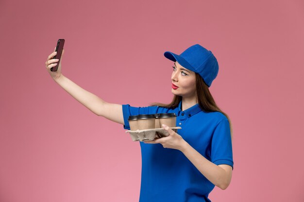 Vue de face femme courrier en uniforme bleu et cape tenant des tasses à café en prenant une photo sur le mur rose