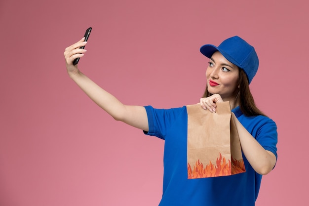 Vue de face femme courrier en uniforme bleu et cape tenant un téléphone et un paquet alimentaire prenant le téléphone sur le plancher rose