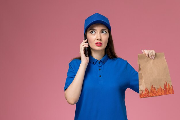 Vue de face femme courrier en uniforme bleu et cape tenant un téléphone et un paquet de nourriture sur le mur rose