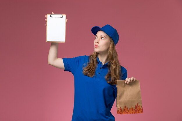 Vue de face femme courrier en uniforme bleu tenant le bloc-notes et l'emballage alimentaire sur le bureau rose entreprise uniforme de service des travailleurs