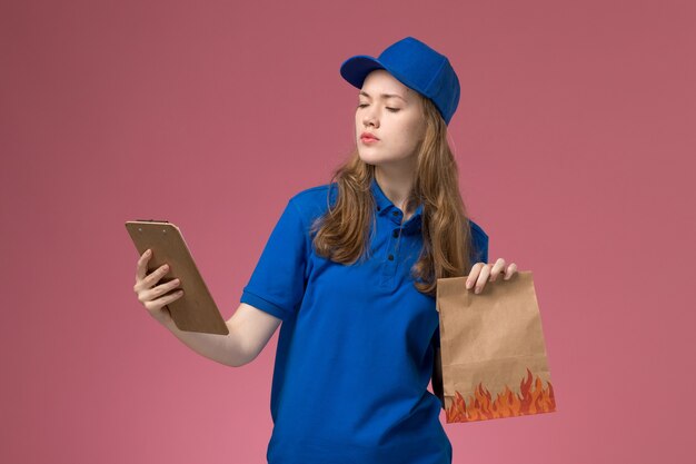 Vue de face femme courrier en uniforme bleu tenant le bloc-notes et le paquet de nourriture sur le bureau rose clair entreprise uniforme de service des travailleurs de l'emploi
