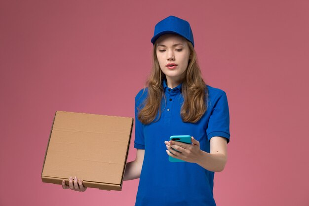 Vue de face femme courrier en uniforme bleu tenant la boîte de livraison de nourriture et à l'aide de téléphone sur le travail de l'entreprise uniforme de service de bureau rose