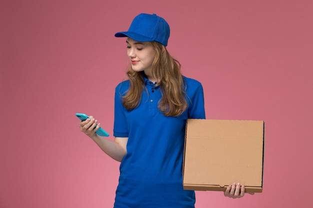 Vue de face femme courrier en uniforme bleu tenant la boîte de livraison de nourriture et le téléphone sur le travail de l'entreprise uniforme de bureau rose