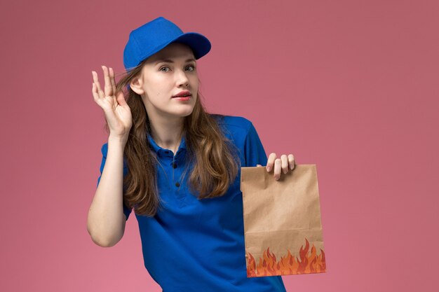 Vue de face femme courrier en uniforme bleu tenant le paquet alimentaire essayant d'entendre sur le bureau rose entreprise uniforme de service des travailleurs