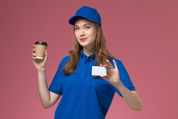 Vue de face femme courrier en uniforme bleu tenant une tasse de café marron avec carte blanche sur un travail uniforme de travail de bureau rose clair entreprise de livraison