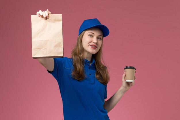 Vue de face femme courrier en uniforme bleu tenant une tasse de café marron avec un paquet de nourriture sur un uniforme de travail de bureau rose clair entreprise de livraison