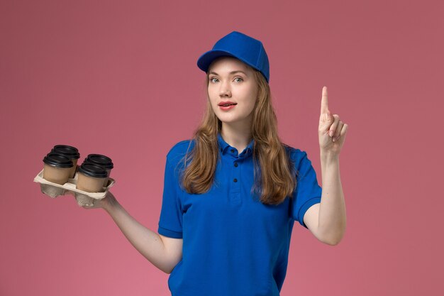 Vue de face femme courrier en uniforme bleu tenant des tasses de café de livraison marron avec le doigt sur le bureau rose des travailleurs de l'entreprise uniforme