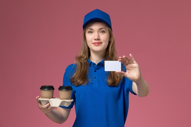 Vue de face femme courrier en uniforme bleu tenant des tasses de café de livraison marron souriant tenant une carte blanche sur l'uniforme de service de bureau rose entreprise de livraison