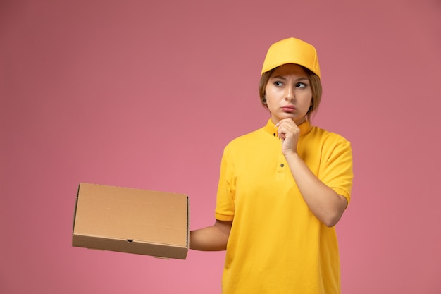 Vue de face femme courrier en uniforme jaune cape jaune tenant la boîte de nourriture pensant sur le bureau rose couleur féminine de livraison uniforme