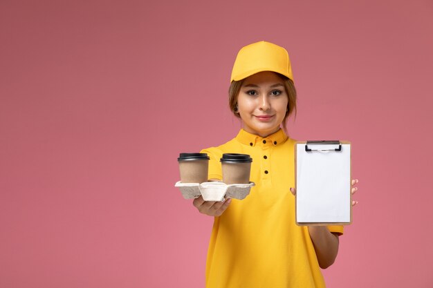 Vue de face femme courrier en uniforme jaune cape jaune tenant café et bloc-notes sur fond rose travail de livraison uniforme