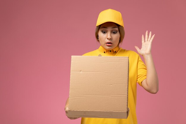 Vue de face femme courrier en uniforme jaune cape jaune tenant et ouvrant la boîte de nourriture sur le bureau rose couleur féminine de livraison uniforme