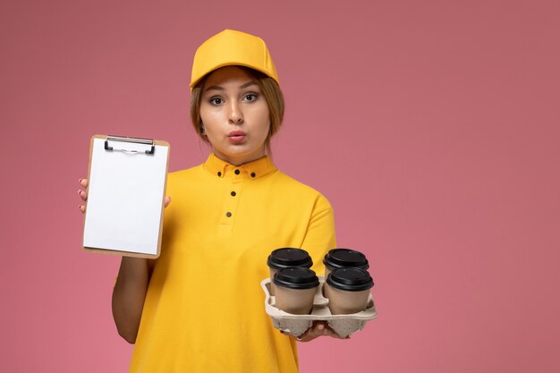 Vue de face femme courrier en uniforme jaune cape jaune tenant des tasses à café et bloc-notes blanc sur fond rose travail de livraison uniforme travail couleur