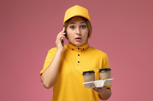 Vue de face femme courrier en uniforme jaune cape jaune tenant des tasses à café et parler au téléphone sur le travail de couleur de travail de livraison uniforme de bureau rose