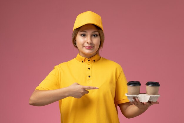 Vue de face femme courrier en uniforme jaune cape jaune tenant des tasses à café en plastique avec sourire sur le travail de livraison uniforme fond rose