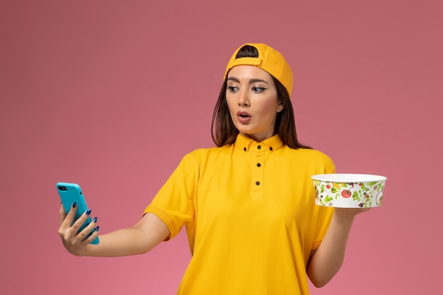 Photo gratuite vue de face femme courrier en uniforme jaune et cape tenant le bol de livraison et à l'aide d'un téléphone sur la livraison uniforme de service de mur rose clair