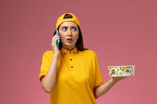 Vue de face femme courrier en uniforme jaune et cape tenant le bol de livraison et à l'aide d'un téléphone parlant sur le travail de livraison uniforme de service de mur rose clair